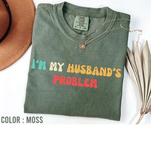 Könnte beinhalten: Moosgrünes T-Shirt mit dem Text "I'M MY HUSBAND'S PROBLEM" in Retro-Schrift. Das Shirt hat einen Rundhalsausschnitt und kurze Ärmel. Eine goldfarbene Halskette mit Anhänger ist auf dem Shirt drapiert. Das Shirt ist aus einem weichen, bequemen Material.