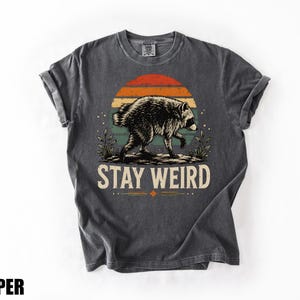 K&ouml;nnte beinhalten: Ein anthrazitfarbenes Kurzarm-T-Shirt mit einer Grafik eines Waschb&auml;ren, der vor einem Sonnenuntergang mit horizontalen Streifen in Orange, Gelb und Blaugr&uuml;n l&auml;uft. Der Text "STAY WEIRD" ist in cremefarbener Schrift unter der Grafik gedruckt.