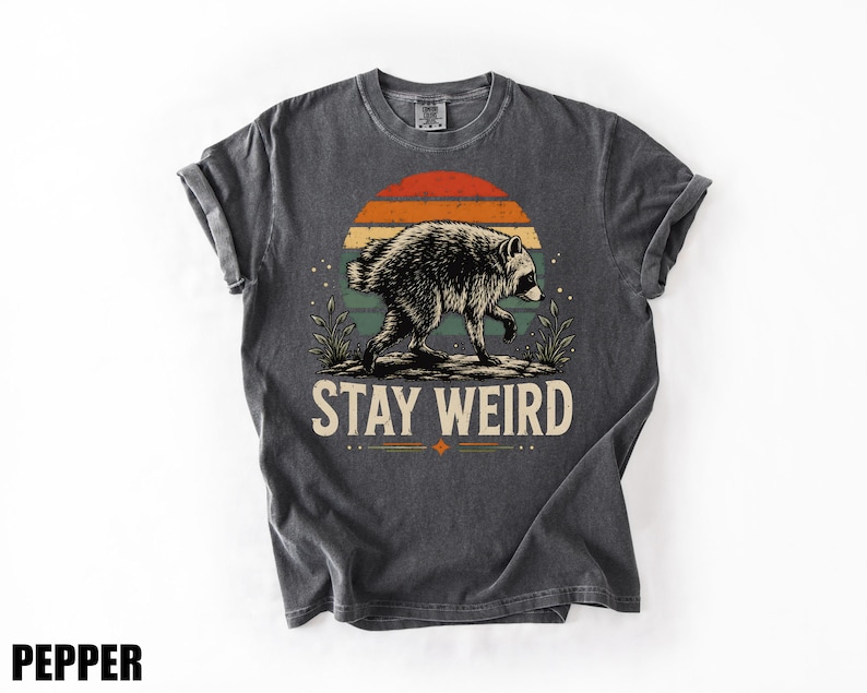 K&ouml;nnte beinhalten: Ein anthrazitfarbenes Kurzarm-T-Shirt mit einer Grafik eines Waschb&auml;ren, der vor einem Sonnenuntergang mit horizontalen Streifen in Orange, Gelb und Blaugr&uuml;n l&auml;uft. Der Text "STAY WEIRD" ist in cremefarbener Schrift unter der Grafik gedruckt.