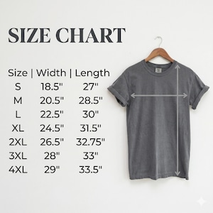 K&ouml;nnte beinhalten: Ein anthrazitfarbenes T-Shirt an einem Holzkleiderb&uuml;gel neben einer Gr&ouml;&szlig;entabelle. Die Tabelle listet die Gr&ouml;&szlig;en S bis 4XL mit den entsprechenden Breiten- und L&auml;ngenma&szlig;en in Zoll auf, von 18,5 mal 27 Zoll bis hin zu 29 mal 33,5 Zoll.
