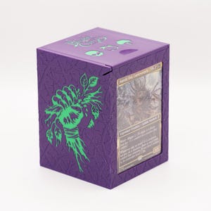 Puede incluir: Una caja morada para guardar cartas con un gráfico verde de una mano sosteniendo plantas. La caja tiene el texto "Blight Crew" y gráficos de calaveras en la tapa. Se ve una carta con el texto "Auntie Dot, Curseweaver".