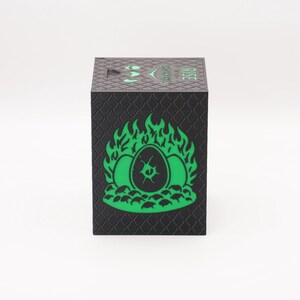 May include: Black cube-shaped box with a textured, scale-like pattern. The front features a green graphic of three eggs engulfed in flames. The word "RISE" is printed in green on the top. A logo is also visible.
