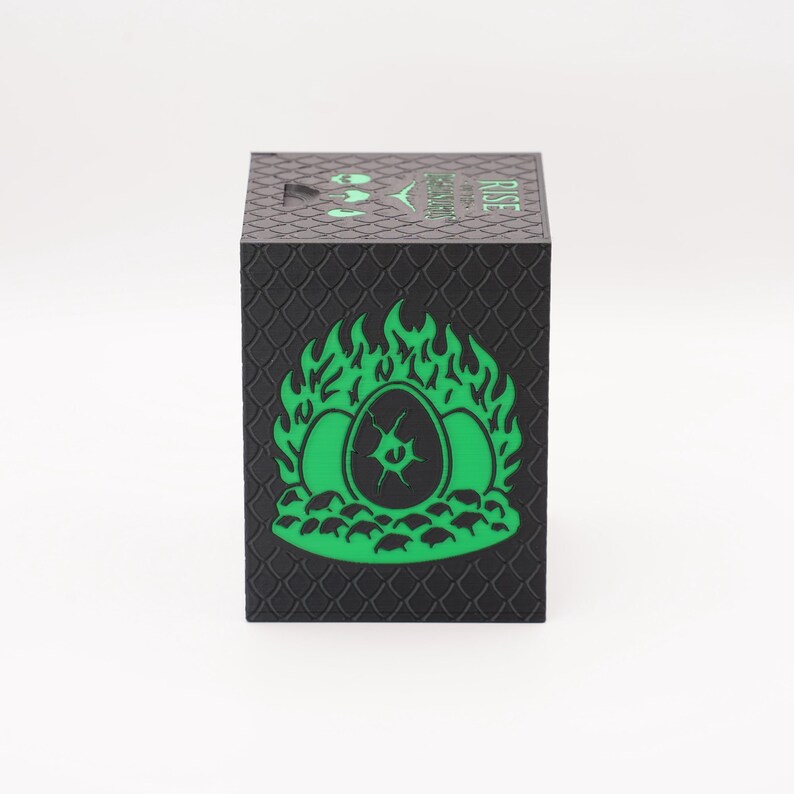 May include: Black cube-shaped box with a textured, scale-like pattern. The front features a green graphic of three eggs engulfed in flames. The word "RISE" is printed in green on the top. A logo is also visible.
