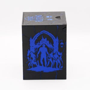 May include: Black cube-shaped box with a blue graphic design. The design features a silhouette of a figure in front of a building, with other figures around it. The top of the box has the text "RISE OF THE PUTTING".