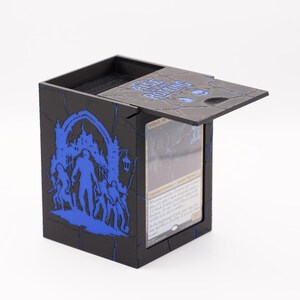May include: Black card storage box with a hinged lid, featuring blue graphics of a castle, figures, and text. The lid is slightly open, revealing a card inside. The box has a textured, cracked appearance.