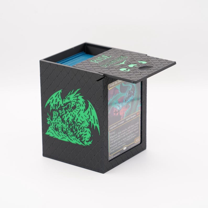 May include: Black card storage box with a hinged lid, featuring a green dragon graphic on the side. The lid has the text "RISE" and other text in green. The box is open, revealing blue cards and a card with a dragon illustration.