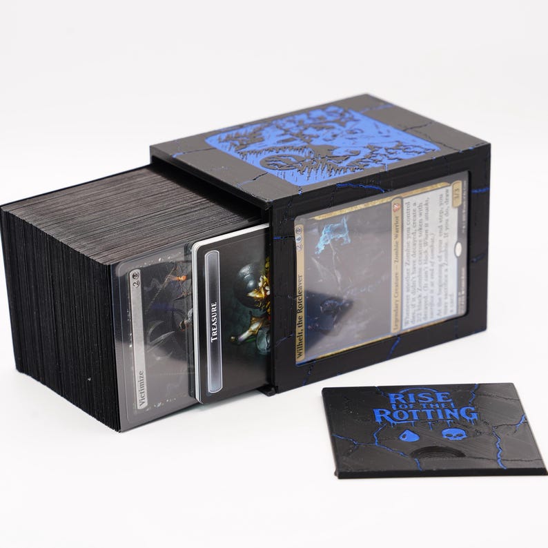 May include: Black wooden card storage box with blue accents, holding a deck of cards. The box features a sliding drawer and a separate lid. The cards are visible, with the text "Victimize" and "Treasure" on the card faces. A small card with the text "RISE FOR THE ROTTING" is also present.
