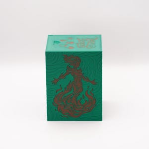 May include: A green box with a detailed design. The box features a brown illustration of a woman engulfed in flames. The top of the box has Chinese characters and other symbols. The box is a cube shape.
