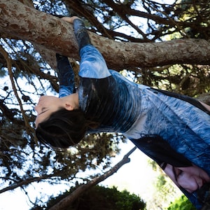 May include: A person wearing a blue and white patterned long-sleeved shirt, hanging upside down from a tree branch.