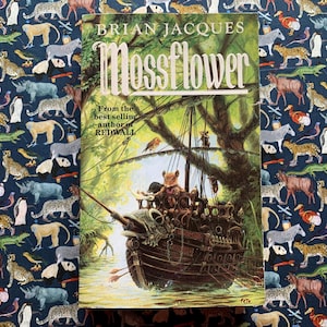Könnte beinhalten: Das Buch "Mossflower" von Brian Jacques mit einer detaillierten Illustration eines Schiffes mit Tierfiguren in einer Waldszene. Das Buch liegt auf einem Stoff mit Tiermotiven.