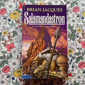 Könnte beinhalten: Ein Buch mit dem Titel "Salamandastron" von Brian Jacques. Das Cover zeigt einen Dachs in goldener Rüstung, der einen Speer hält, vor einem Berg- und Sonnenuntergangshintergrund. Das Buch liegt auf einem Stoff mit Blumenmuster.
