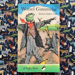 Puede incluir: Una copia vintage de "Worzel Gummidge" de Barbara Euphan Todd, un libro Puffin. La portada presenta una ilustración de Worzel Gummidge, un espantapájaros, con una cabeza verde, una bufanda roja y un abrigo. El libro está sobre un fondo azul oscuro.
