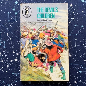 May include: A vintage paperback book titled "The Devil's Children" by Peter Dickinson. The cover features a colorful illustration of a group of people in a snowy landscape, with a Penguin Books logo in the upper left corner.