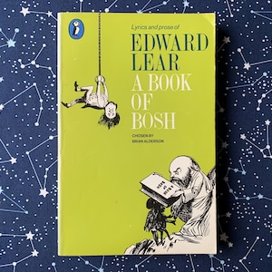 Puede incluir: Un libro de bolsillo vintage titulado "Edward Lear: A Book of Bosh" con una cubierta verde lima. La portada presenta ilustraciones de una persona colgada de una cuerda y un hombre barbudo leyendo un libro. El fondo es una tela azul oscuro con un patrón de constelación blanca.