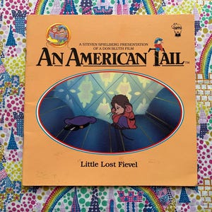 May include: An orange book cover with the title "An American Tail" and the words "Little Lost Fievel." The cover features an illustration of a mouse in a red coat and a purple hat.