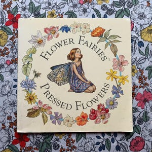 May include: A vintage book cover titled "Flower Fairies Pressed Flowers" features a seated fairy surrounded by a floral wreath. The cover has a cream background with colourful flowers and text. The fairy has butterfly wings and is wearing a blue dress.