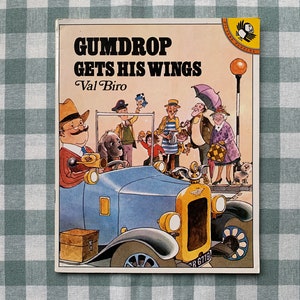 May include: A vintage children's book cover titled "Gumdrop Gets His Wings" by Val Biro. The illustration depicts a blue car with a dog driving, surrounded by cartoon figures. The background is a green and white checkered pattern.