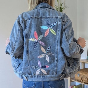 Op de afbeelding: Een spijkerjack met een bloemenpatroon op de rug. Het ontwerp is gemaakt van meerdere stofresten in verschillende kleuren en patronen.