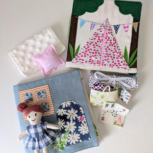 May include: A collection of handmade fabric toys, including a doll, a tent-shaped book, and a small pillow. The tent book features a floral pattern and bunting. The doll wears a blue and white checkered dress. Other items include a small basket and a map.