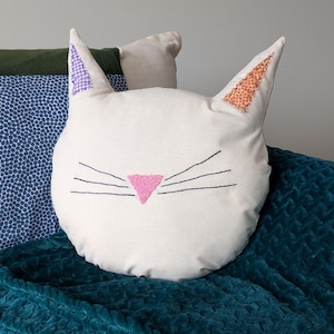 Op de afbeelding: Een wit kussenvormig kussen in de vorm van een kat met geborduurde zwarte snorharen en een roze driehoekige neus. De oren hebben stoffen met paarse en oranje patronen. Het kussen ligt op een turquoise deken en andere kussens.