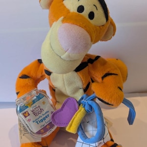 May include: A plush Tigger toy from Winnie the Pooh, with orange fur and black stripes, a pink nose, and a tag. The toy holds a purple heart, a yellow rectangle, and a blue and white patterned object.