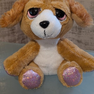 Könnte beinhalten: Ein Plüschhundespielzeug mit braunem Fell, Schlappohren und einer weißen Brust. Das Spielzeug hat große, schwarze Augen mit rosa Akzenten und glitzernden lila Pfoten. Der Hund hat einen traurigen Gesichtsausdruck.