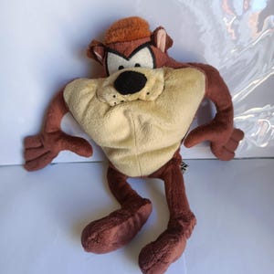 Może przedstawiać: Pluszowa zabawka przedstawiająca Tasmańskiego Diabła, postać z kreskówki. Zabawka ma beżowy brzuch, brązowe ramiona i nogi oraz brązową głowę z małym kapeluszem. Twarz ma białe, czarne i brązowe detale.