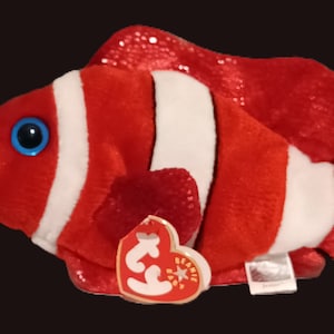 May include: A plush clownfish toy with red and white stripes, blue eyes, and a blue nose. The fish has a tag attached. The fins and tail have a glittery texture. The toy is designed to resemble a clownfish.
