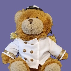 Könnte beinhalten: Ein brauner Teddybär mit einer weißen Matrosenjacke mit goldenen Verzierungen und Messingknöpfen sowie einer passenden Matrosenmütze. Der Bär hat eine hellbraune Schnauze, vor einem violetten Hintergrund.