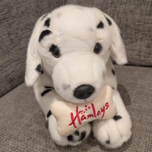 May include: A plush Dalmatian dog with black spots on a white body. The dog is holding a bone-shaped toy with the embroidered text "Hamleys" in red. The toy dog has black eyes and a black nose.