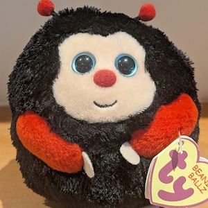 May include: A soft ladybird toy with a black body, red arms, and red antennae. The toy has large blue eyes, a red nose, and a smiling face. A tag that says "BEANIE BALLZ" is attached to the toy.