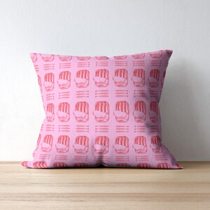 May include: A square pink cushion featuring a repeating pattern of red abstract shapes and horizontal lines. The cushion is on a wooden surface against a white wall.