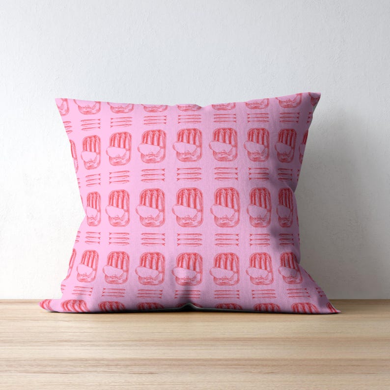 May include: A square pink cushion featuring a repeating pattern of red abstract shapes and horizontal lines. The cushion is on a wooden surface against a white wall.