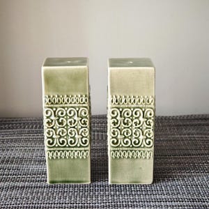 May include: Two tall, rectangular ceramic salt and pepper shakers in a light sage green glaze. Each shaker features a central band of intricate, raised scrollwork patterns and smaller decorative borders at the top and bottom of the textured section.