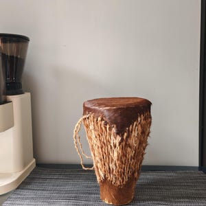 May include: A djembe drum featuring a dark brown animal hide head and a body wrapped in light tan, fibrous material. The drum has a tapered cylindrical shape with a textured, hairy surface and a braided rope handle attached to the side.