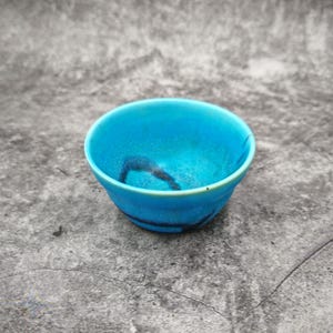 May include: A small, round ceramic bowl featuring a vibrant turquoise blue glaze with a smooth, glossy finish. The interior and exterior surfaces display subtle, dark, organic-shaped markings that contrast against the bright blue colour.