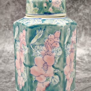 May include: A hexagonal ceramic jar with a lid. The jar is teal with pink floral designs. The lid also has a floral pattern. The jar has a glossy finish and a cream-coloured base.