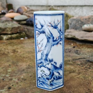 May include: A rectangular porcelain vase featuring a blue and white tree and bird design. The vase has a geometric shape with a slightly flared top and a detailed illustration of a tree with small pink blossoms.