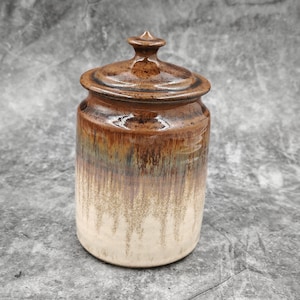 May include: A ceramic jar with a lid. The jar is cylindrical with a rounded top and a small knob on the lid. The colors transition from brown to beige, with a dripped glaze effect. The jar is likely handmade.