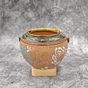 May include: A round ceramic pot with a textured tan surface featuring embossed floral patterns in white and turquoise. The rim is glazed in a dark green and brown finish with a beaded detail. The pot rests on a small, light-coloured wooden block base.