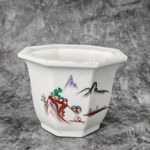Könnte beinhalten: Weißer Keramik-Blumentopf mit achteckiger Form. Der Topf zeigt eine handgemalte Landschaftsszene mit Bergen, Bäumen und Figuren in Rot, Grün, Lila und Schwarz. Geeignet für kleine Pflanzen.