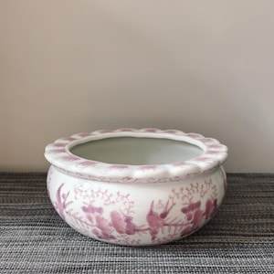 May include: A white ceramic bowl with a scalloped rim and a pink floral pattern painted around the exterior. The bowl features a rounded body and a smooth, glossy finish, with delicate pink vines and blossoms decorating the curved surface.