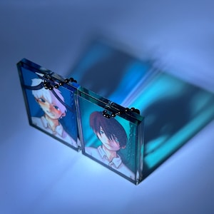 May include: Two clear acrylic panels hinged together, each displaying a colorful anime-style illustration of a person. The panels are connected by small black hinges. The background is a gradient of blue.