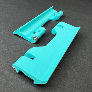 May include: Two turquoise-colored, plastic components with curved edges and metal screws. One component is hinged, and the other has a curved notch. The items are set against a dark gray background.