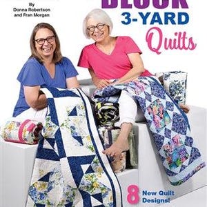 May include: A book cover titled "One Block 3-Yard Quilts" featuring two women and a quilt. The quilt is white with blue and yellow floral patterns. The cover also includes the text "8 New Quilt Designs!" and the Fabric Cafe logo.