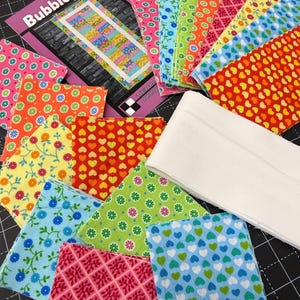 Può includere: Una collezione di quadrati e strisce di tessuto colorati con motivi floreali, a cuore e geometrici. I tessuti sono in tonalità di rosa, blu, verde, arancione e giallo. È presente anche un libretto di modelli intitolato "Bubblegum".