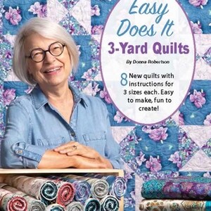 Könnte beinhalten: Eine Frau lächelt vor einem floralen Quilt. Das Bild zeigt ein Buch mit dem Titel "Easy Does It 3-Yard Quilts" von Donna Robertson. Das Buchcover zeigt ein florales Quiltmuster. Gerollte Stoffballen befinden sich in einem Holzregal.