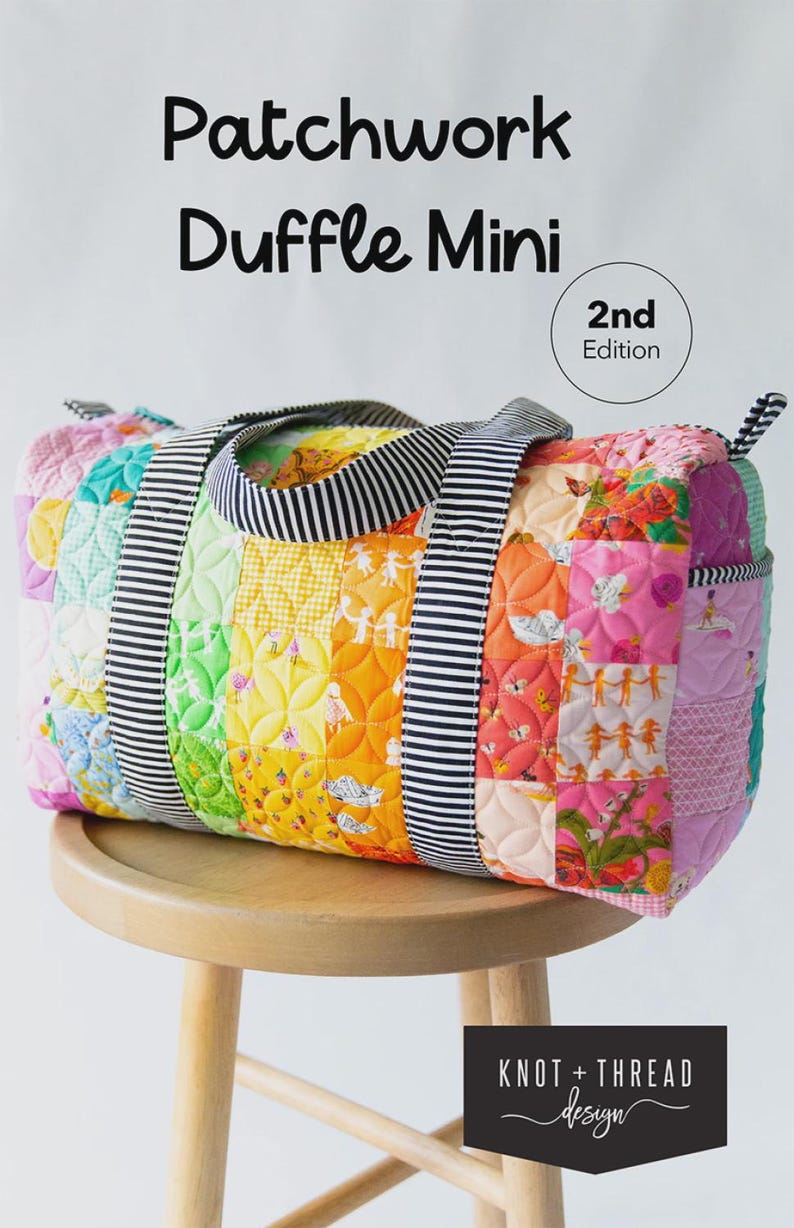 May include: A vibrant patchwork duffle bag with a striped handle. The bag features a variety of patterned fabrics in a rainbow of colours. It is labelled "Patchwork Duffle Mini" and "2nd Edition". The bag is displayed on a wooden stool.