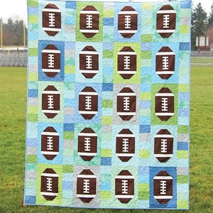 May include: A patchwork quilt featuring brown footballs on a blue, green, and gray background. The word "Touchdown" is at the top. The quilt is displayed on a grassy field.