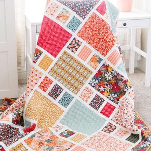 May include: A quilt with a geometric pattern of squares and rectangles in shades of red, orange, yellow, blue, and green. The quilt is made with a variety of floral fabrics. The quilt is folded and displayed on a white table with a white drawer. The text "Craftsman" is displayed in a blue banner at the top of the image. The text "Amy Smart Diary of a Quilter" is displayed in a white banner at the bottom of the image.